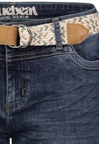Geflochtener Stoffgürtel mit Lederenden und Metallringen, getragen zu dunkelblauen Jeans mit sichtbarem Markenetikett im Innenbund.