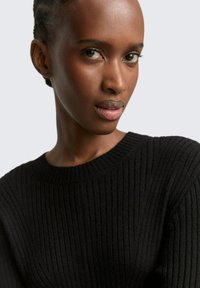 Schwarzer gerippter Pullover mit rundem Halsausschnitt und langen Ärmeln, vor einem neutralen Hintergrund präsentiert. Weicher Stoff und figurbetontes Design.