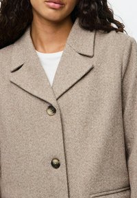 Beige villasegu segu coat, millel on tekstuuriga herringbone muster, lõhkega krae ja kaks nuppu. Omab küljetaskuid ja lõdva istuvust.