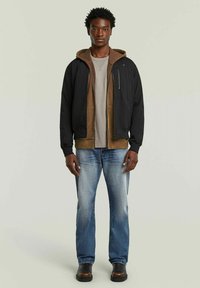 Chaqueta bomber negra en capas con acentos marrones, camiseta gris, vaqueros azules y botas oscuras, sobre un modelo frente a un fondo verde claro.