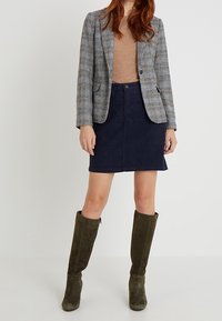 Blazer à carreaux gris et bleu, haut beige, jupe en daim bleu foncé, bottes vert olive hauteur genou, mettant en avant une coupe ajustée et un design structuré.