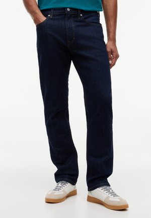 Moški, oblečen v temno modre kavbojke iz džinsa in bele superge z rjavkastimi podplati, stoji z eno roko v žepu pred belim ozadjem.