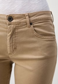 Gros plan d'une personne portant un pantalon beige décontracté avec un bouton noir et une poche avant, associé à un haut blanc texturé.