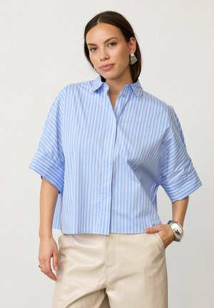 Kobieta w niebiesko-białej paskowanej koszulce z krótkim rękawem, beżowych spodniach, srebrnej bransoletce i kolczykach, pozująca z jedną ręką w kieszeni.