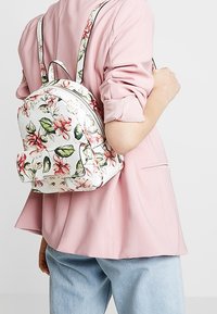 Rugzak met bloemenpatroon, witte basis, leerachtige buitenkant, gebogen vorm en verstelbare bandjes, vastgehouden tegen een pastelroze kledingstuk.