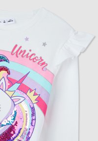 Biała koszulka z krótkim rękawem z grafiką jednorożca, wielokolorowymi paskami, cekinami i falbankami na rękawach. Nad wzorem znajdują się gwiazdki.