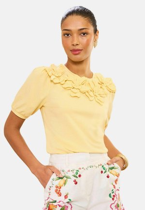 Žena v žltom topu s kvetinovými aplikáciami a bielych nohaviciach s farebnou ovocnou a kvetinovou výšivkou, pózujúca s rukami v vreckách.