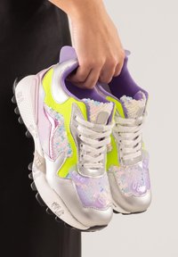 Una mano sostiene un par de zapatillas plateadas, moradas y amarillas neón con lentejuelas y cordones blancos sobre un fondo neutro.