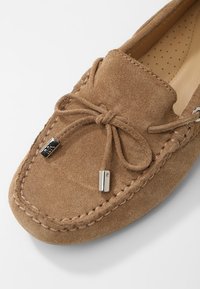 Mocassin en suede marron avec un bout arrondi, lacets décoratifs, accents en métal et détails cousus autour de la semelle. Doublure intérieure lisse.
