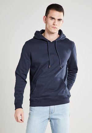 Navyfarbener Baumwoll-Hoodie mit einer Fronttasche, verstellbaren Kordeln und gerippten Bündchen. Modell trägt hellblaue Jeans.