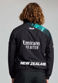 Giubbotto a vento nero con un design a gradiente turchese, con la scritta "Emirates Fly Better" e "Nuova Zelanda" sul retro. Tessuto leggero.