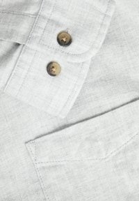 Tissu gris en gros plan montrant deux boutons marron et une couture détaillée autour d'une poche et d'un rabat boutonné.