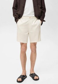Beige shorts lavet af letvægtsstof, med knaplukning og sidelommer, båret med sorte sandaler.