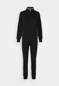 Chaqueta negra con cremallera y un cuello alto, bolsillos laterales con cremallera, combinada con pantalones de chándal negros, ambos hechos de un tejido suave y texturizado.