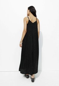 Robe maxi plissée noire avec des bretelles fines, affichant une texture scintillante. Portée avec des chaussures noires à motifs, vue de dos.