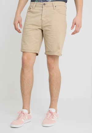 Man in beige opgerolde shorts en roze sneakers met witte strepen, staande tegen een effen lichte achtergrond.