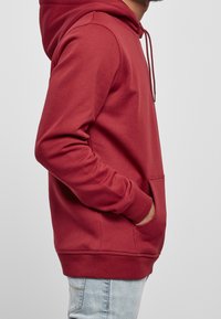 Sweat à capuche rouge avec poche avant, capuche à cordon, poignets et ourlet côtelés, en tissu doux. Porté avec un jean bleu clair.