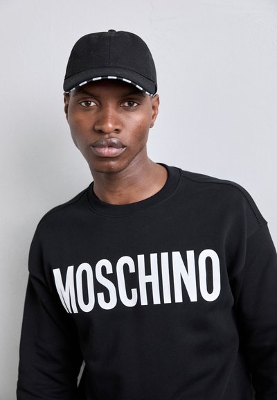 Sudadera negra con "MOSCHINO" en grandes letras blancas. Cuenta con un cuello redondo y costuras visibles. Se lleva con una gorra negra con un visor a rayas.