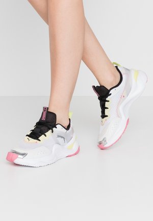 Piernas con zapatillas Puma blancas, rosas, negras y amarillas, de pie sobre un suelo gris claro con los tobillos cruzados.