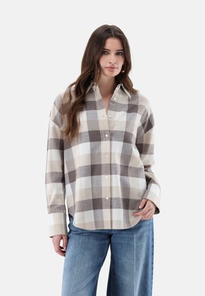 Junge Frau mit langen Haaren, die ein beige-grau kariertes Hemd mit Knöpfen und blaue Jeans trägt, steht vor einem einfarbigen hellen Hintergrund.