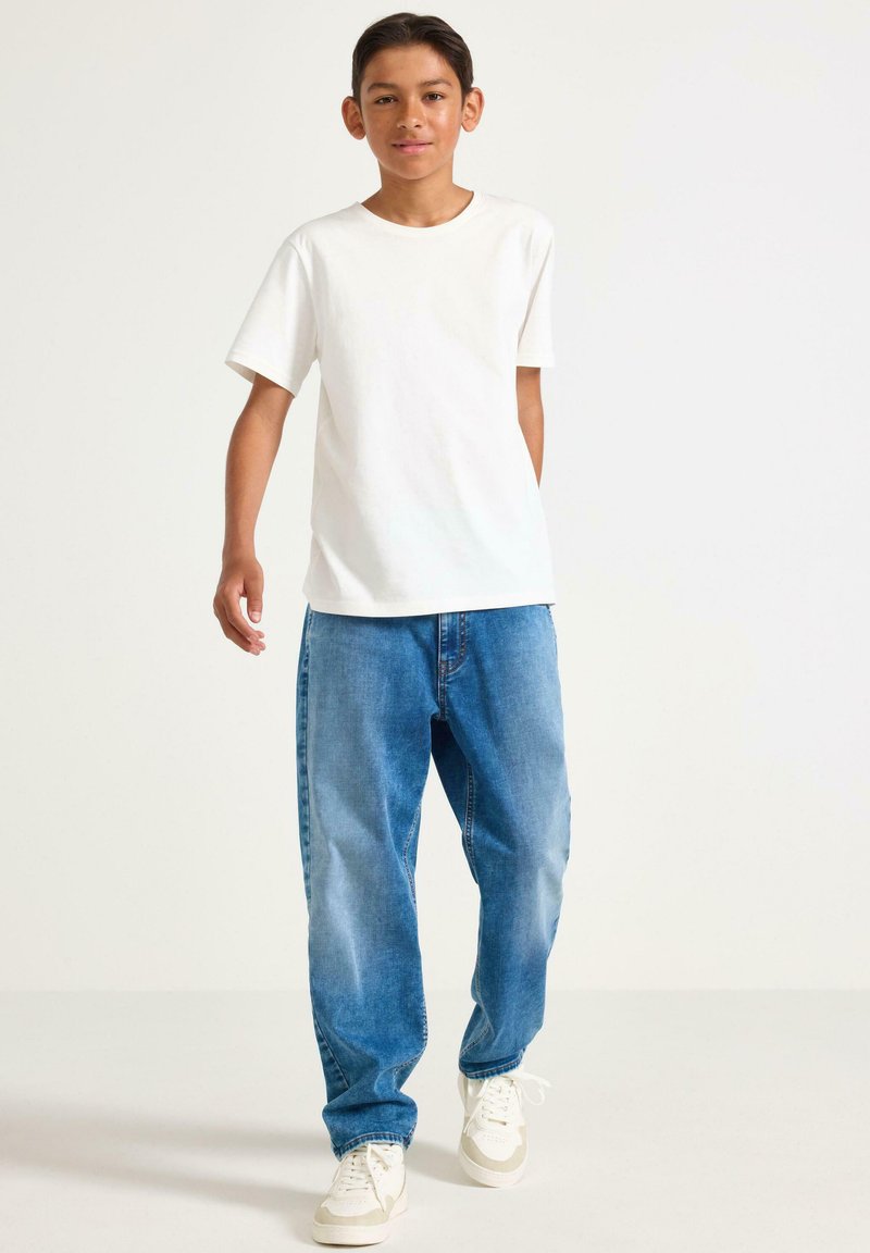 Hvid bomulds t-shirt, løstsiddende blå denim jeans og beige sneakers. Personen står med den ene hånd i lommen, mod en hvid baggrund.