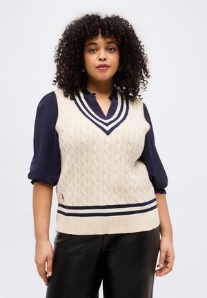 Γυναίκα με σγουρά μαλλιά που φοράει ένα εκρού πουλόβερ αμάνικο με πλέξη καλώδιο και ναυτικές ρίγες πάνω από ένα ναυτικό μπλουζάκι και μαύρο δερμάτινο παντελόνι, όρθια.