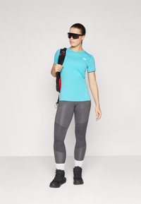 Kék sportpóló, szürke mintás leggings, fekete túracipő és fekete napszemüveg. A modell egy fekete hátizsákot tart, piros részletekkel.