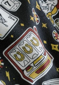 Schwarzer Stoff zeigt ein Muster mit einem roten und gelben Spielautomaten, Würfeln, Spielkarten und Hufeisen, umrandet in kräftigen Farben.