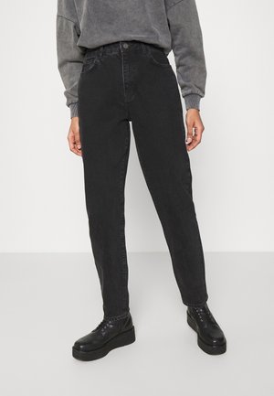 Personne portant un jean noir taille haute, des bottes noires à plateforme avec lacets, et un sweat-shirt gris à manches longues, debout devant un fond uni.