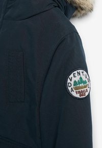 Manche de veste bleu foncé avec capuche bordée de fourrure et écusson circulaire portant l'inscription « Adventure Trails 082 » avec des arbres sur le bras.