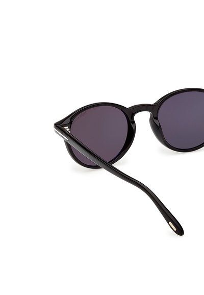 Lunettes de soleil rondes noires avec des verres teintés foncés reposant sur une surface blanche, montrant une branche du côté étendue vers l'avant.