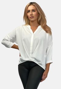Weiße Bluse mit V-Ausschnitt, langen Ärmeln und lockerer Passform. Der Stoff wirkt glatt, mit subtilen Drapierungen am Saum. Kombiniert mit schwarzen Hosen.