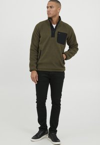 Fleece pullover in olive green with a zip neck, black chest pocket, and side pockets. Paired with black slim-fit jeans and black sneakers.