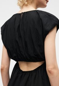 Femme aux cheveux blonds bouclés portant une robe noire avec des manches bouffantes et un dos découpé laissant apparaître la peau entre le haut et la jupe.