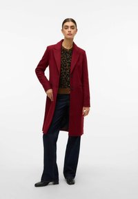 Cappotto in lana bordeaux con vestibilità su misura, rever a punta, due bottoni, tasche a pattina e una trama liscia, indossato sopra una maglietta con stampa leopardata.