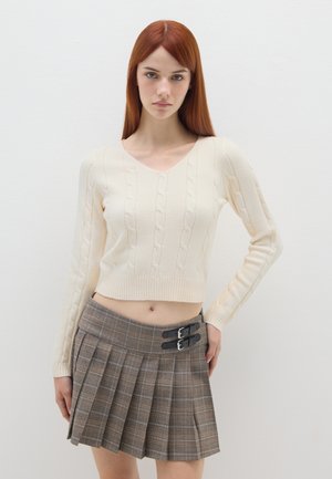 Pullover crema a maglia cable con scollo a V e orlo a costine, abbinato a una gonna a pieghe a quadri marrone con dettaglio a doppia fibbia.