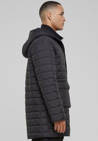 Svart quiltet parkas med hette, med glidelås, brystlomme og sidelommer. Teksturen er glatt med en lett glans.