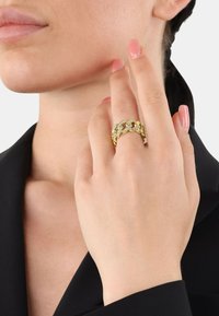 Anello dorato con un design intrecciato, messo in risalto da piccole pietre trasparenti. La mano è leggermente appoggiata sulla guancia, con smalto per unghie delicato.