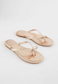 Sandali piatti rosa chiaro con una texture liscia, cinturini sottili e un dettaglio a fiocco al centro. Presentano un design minimalista e una suola piatta.