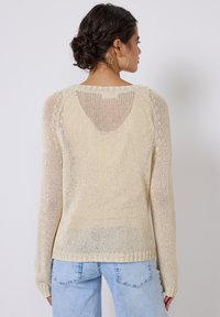 Beige gestrickter Pullover mit langen Ärmeln, weiter Passform und strukturiertem Muster. Der Rücken hat ein leicht offenes Detail am Ausschnitt.