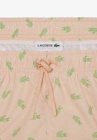 Helle pfirsichfarbene Badeshorts mit grünen Krokodilmustern, elastischem Bund, Kordelzug und weißem Lacoste-Label innen.