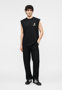 Jeune homme debout portant un débardeur noir avec un logo, un pantalon noir et des baskets noires à rayures blanches, sur un fond uni.