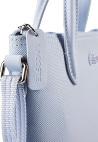 Sac à main Lacoste bleu clair texturé avec une bandoulière amovible et une étiquette de marque en relief attachée près de la fermeture éclair.