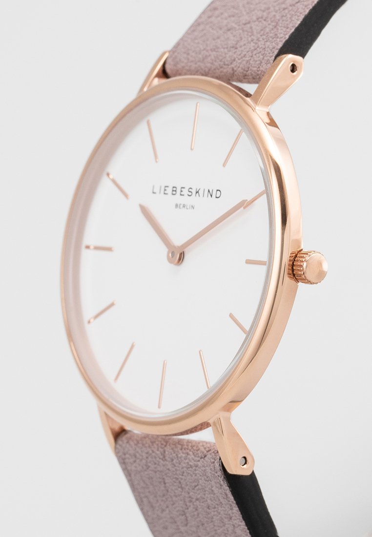Runde Uhr mit weißem Zifferblatt und goldfarbenen Zeigern. Roségold-Gehäuse mit einem pinken Wildlederarmband. Minimalistisches Design mit dünnen Stundenmarkierungen.