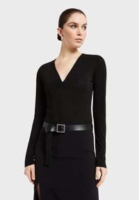 Crna majica dugih rukava s V-izrezom, izrađena od sjajnog materijala, u kombinaciji s crnom suknjom koja ima naglasak u obliku remena s četvrtastom kopčom.