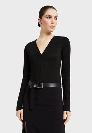 Sort langærmet top med V-hals, lavet af et skinnende materiale, sammen med en sort nederdel, der har et accentbælte med en kvadratisk spænde.