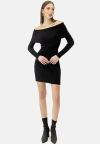 Schwarzes geripptes Off-Shoulder-Kleid mit langen Ärmeln, figurbetonter Passform und mini Länge. Kombiniert mit schwarzen High-Heel-Stiefeletten.