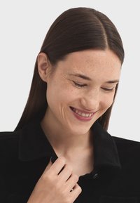 Giovane donna con lunghi capelli castani e lentiggini sorride dolcemente, guardando in basso mentre tiene il colletto della sua giacca nera.