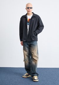 Giacca nera con zip in tessuto testurizzato, t-shirt bianca con grafica, jeans in denim sbiaditi con dettagli strappati e sneaker nere con accenti gialli.