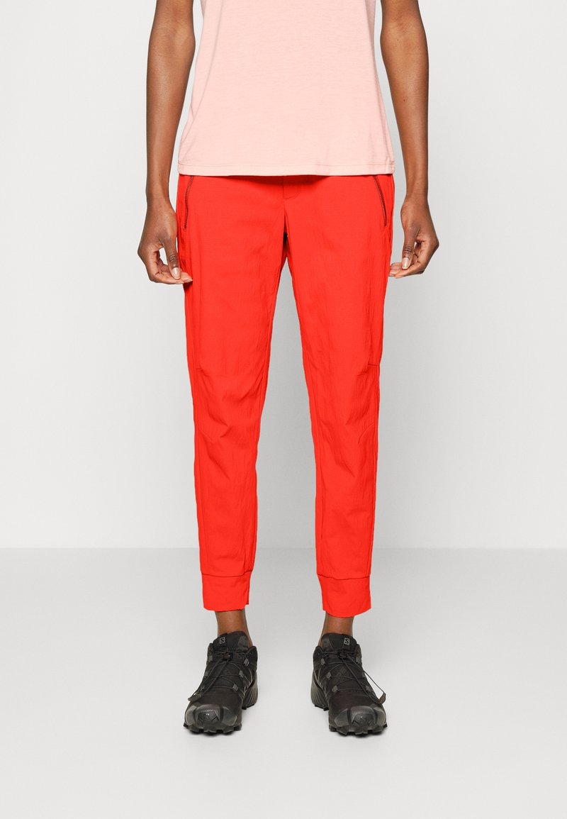 Pantalon de sport rouge avec une finition texturée, poches zippées et poignets ajustés, associé à des chaussures sombres et robustes.